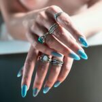 Close-up of hands with long manicured blue nails adorned with silver rings.